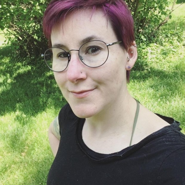 An outdoor photograph of a pale woman with short purple hair, smiling. She is wearing earrings and a black top.