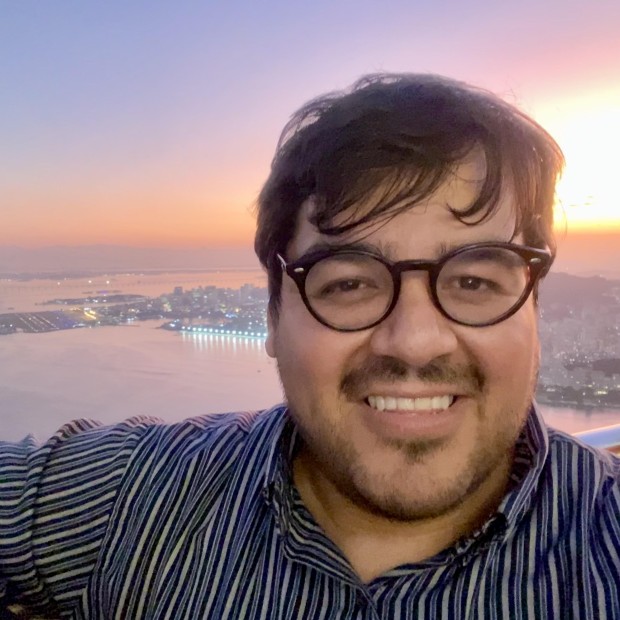Picture of Andy, wearing striped shirt and glasses, standing outside with sunset in background