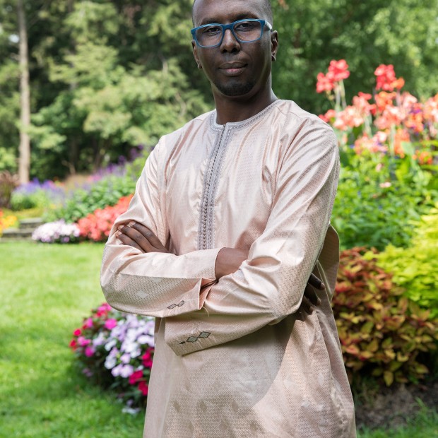 A professional photograph of a man standing in a garden. He wears glasses and has dark skin.