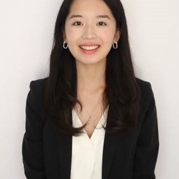A professional photograph of a woman with light skin and dark hair standing in front of a white background. She wears a black suit with a white shirt and is smiling.
