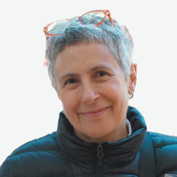 A photograph of a woman with medium-light skin and short gray hair, smiling. She is wearing earrings, a black insulated vest, and glasses on top of her head..