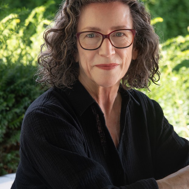 A photograph of a woman with light skin and shoulder-length curly brown hair, smiling. She is wearing red glasses and a black top.
