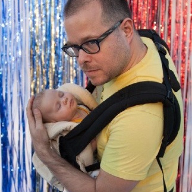 A pale man holds an infant in his arms as they both stand in front of a sparkling background. The man has glasses and wears a yellow shirt.