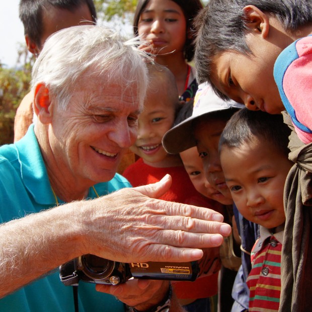 A candid shot of a pale man surrounded by children. He is showing them an image on a camera and smiling.
