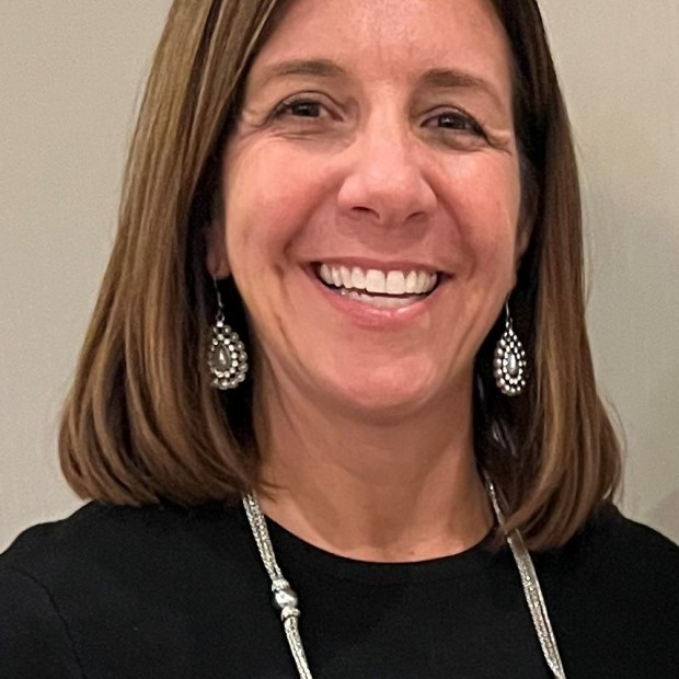 A photograph of a woman with medium-light skin and shoulder length brown hair, smiling widely. She is wearing earrings, a necklace, and a black top.