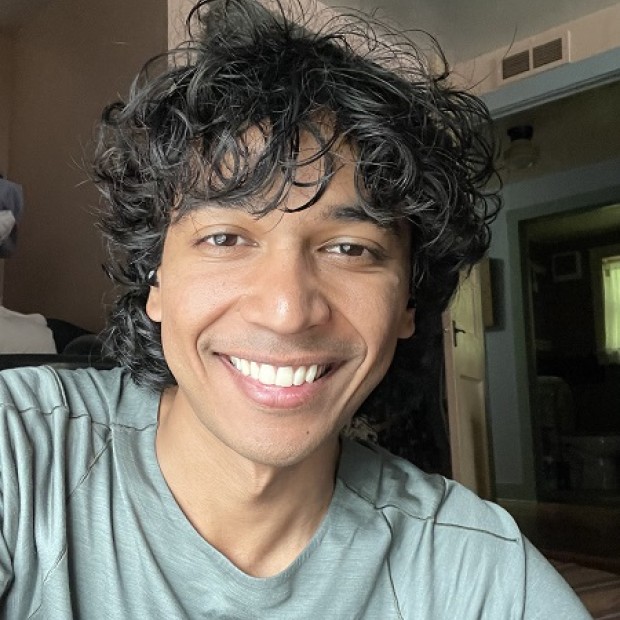A candid shot of a smiling person with medium-brown skin and dark curly hair. They are smiling.
