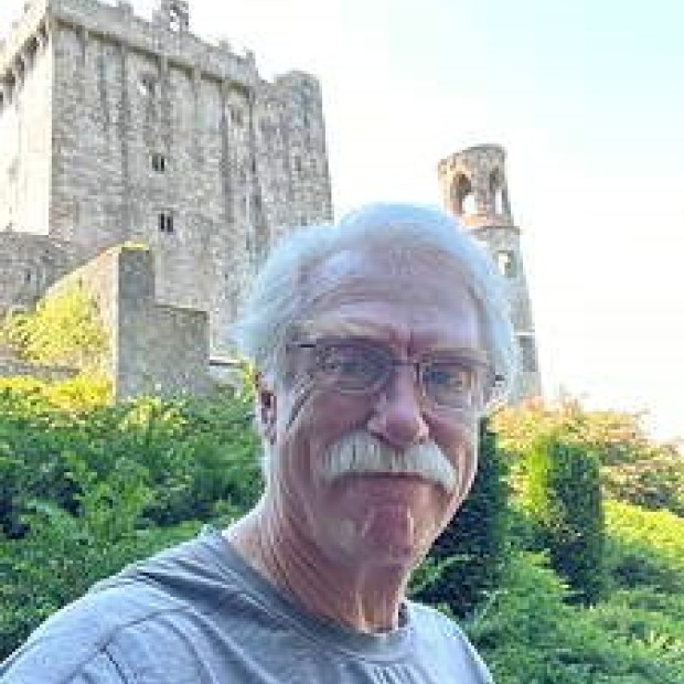 A candid shot of a pale man with a white moustache in front of a stone building.