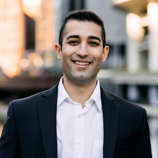 A professional photograph of a smiling man standing in a cityscape. He wears a black sport coat and a white collared shirt.