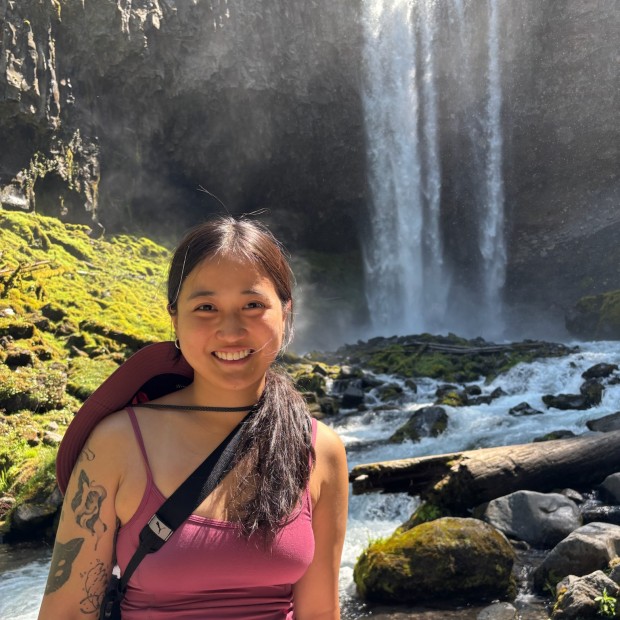 An outdoor photograph of a woman in front of a waterfall. She has light-brown skin, brown long hair, and is smiling.