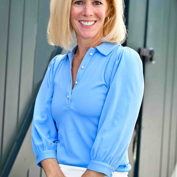 A professional photograph of a long-haired light-skinned woman standing outside. She wears a blue collared shirt and is smiling.