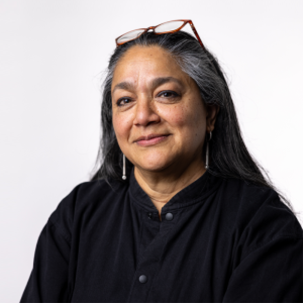 A professional headshot of a woman with medium-brown skin and long dark hair smiling at the camera. She wears a black top and glasses on top of her head.