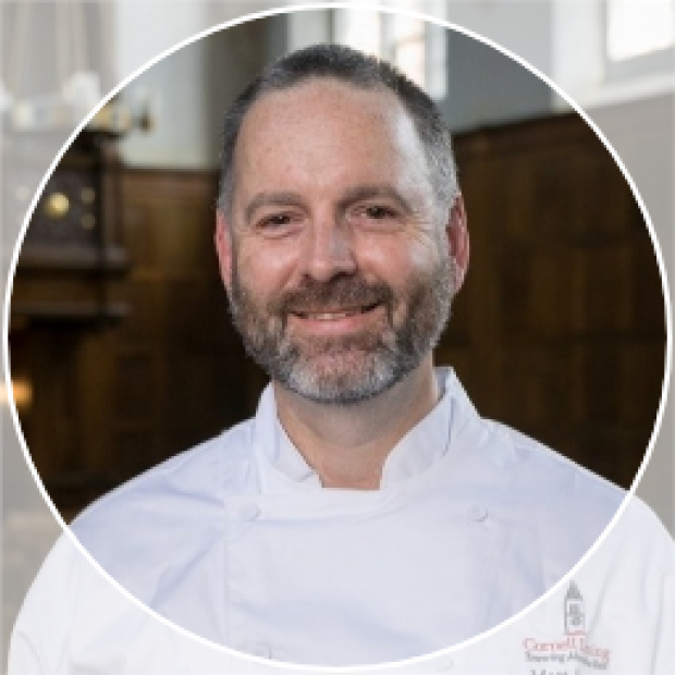 A professional headshot of a man with light skin smiling at the camera. He wears chef's whites and has a short beard.