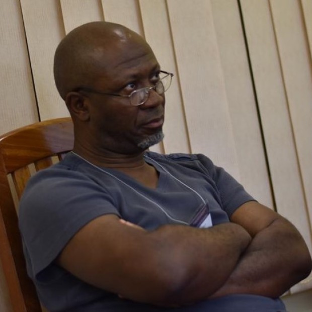 A candid photograph of a seated man. He has dark skin, glasses, and a shaved head. 