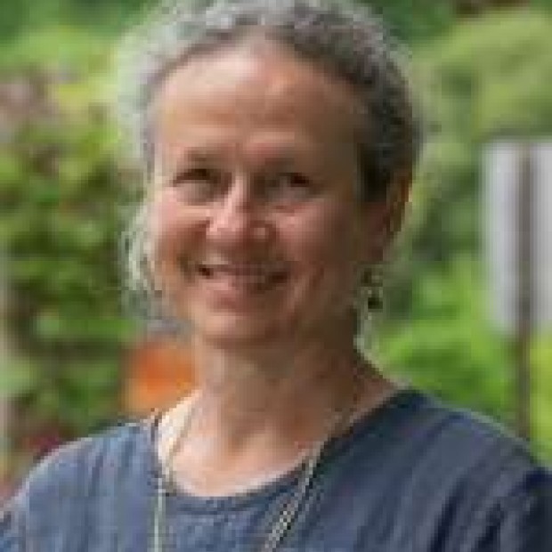 A professional headshot of a woman outdoors smiling at the camera. She wears a blue top and a necklace.