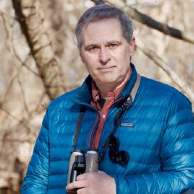 A professional photograph of a light-skinned man outdoors. He has grey short hair, wears binoculars around his neck and has a blue jacket on.