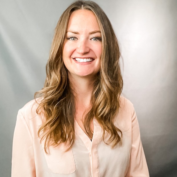 A professional photograph of a long-haired woman seated in front of a gray backdrop. She has medium-light skin and is smiling.