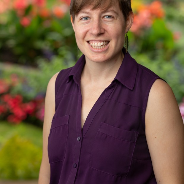A professional photograph of a short-haired woman standing in a garden. She has medium-light skin and is smiling.