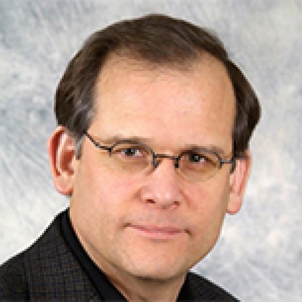 A photograph of a man with light skin and short brown hair. He is wearing glasses and poses in front of a gray background