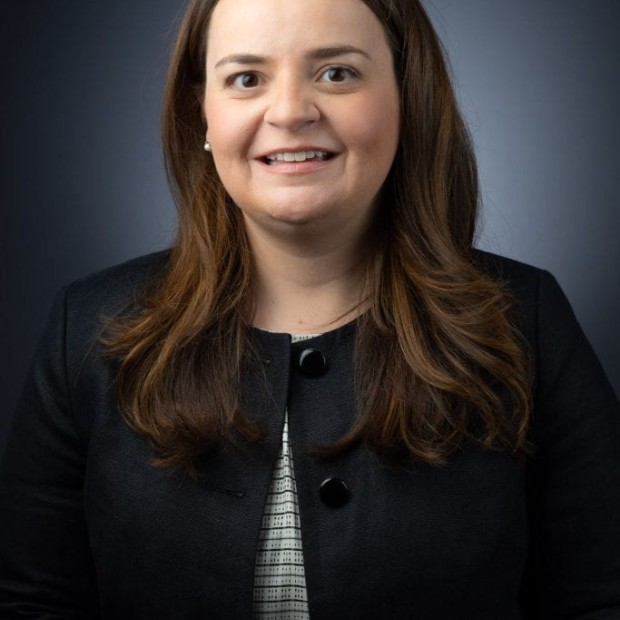 A studio portrait of a light-skinned woman with shoulder length brown hair. She's wearing a dark suit in front of a blue background.