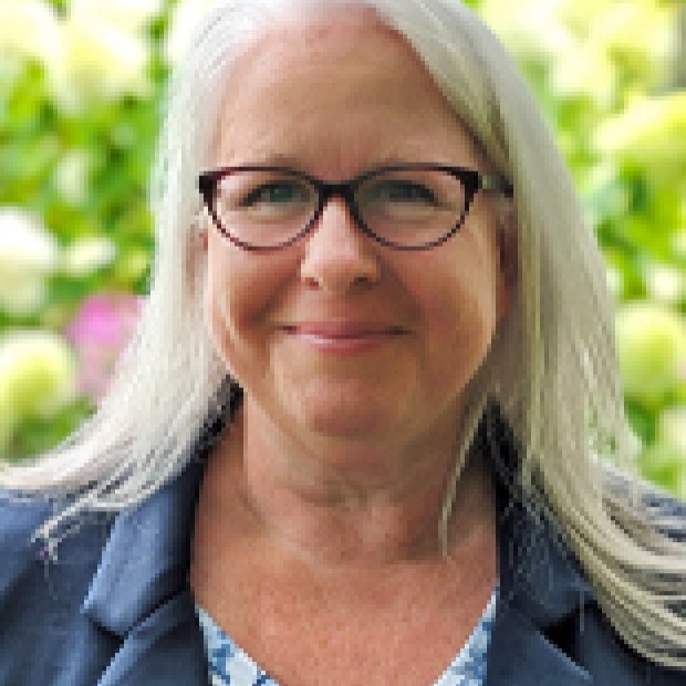 A professional headshot of a woman with pale skin smiling. She wears glasses and a blue jacket.