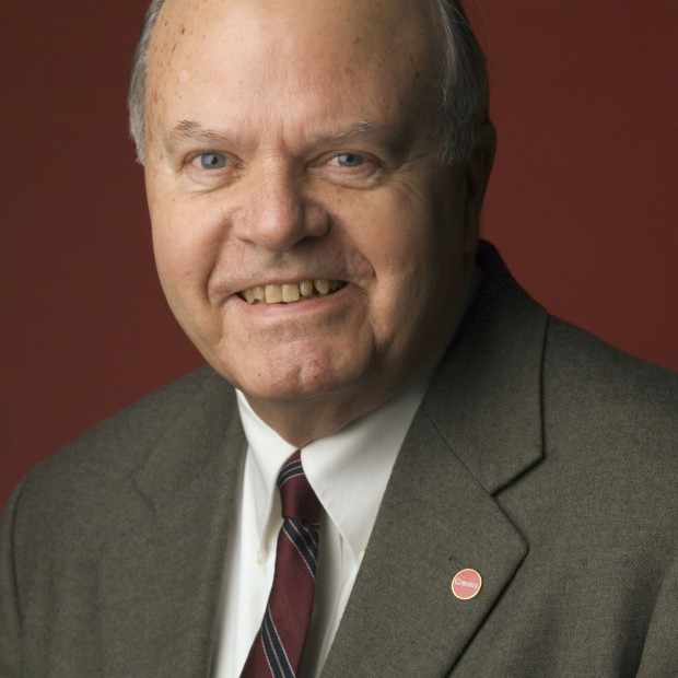 A professional headshot of a man with light skin smiling at the camera. He wears a brown suit and striped tie.
