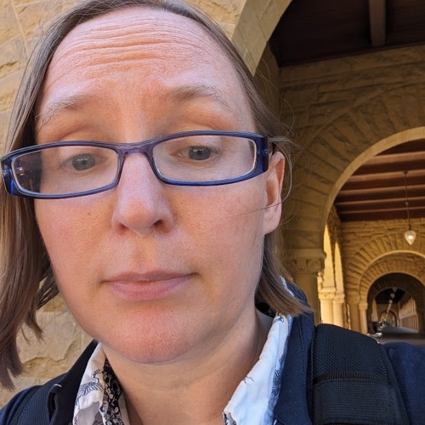 A photograph of a light-skinned woman wearing glasses. She's wearing a collared shirt and jacket and standing in front of a stone building.