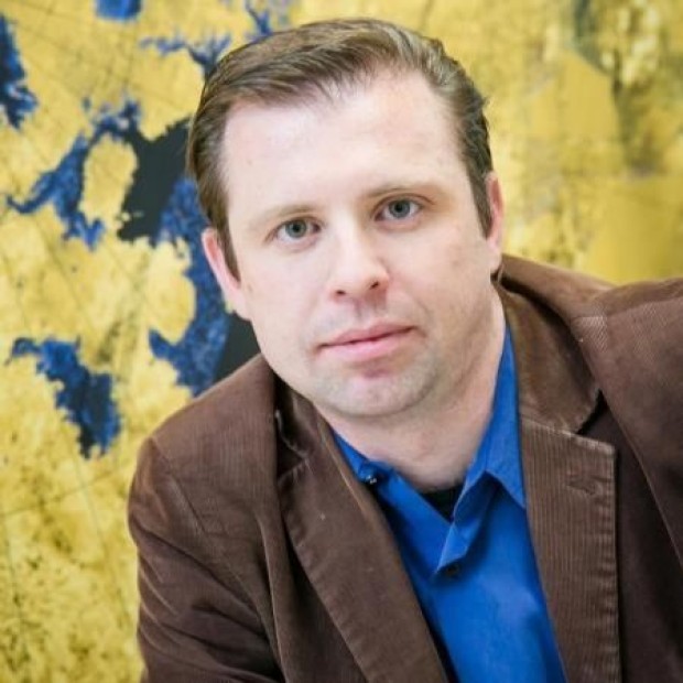 Person in a brown jacket and blue shirt seated in front of a yellow and blue abstract background.