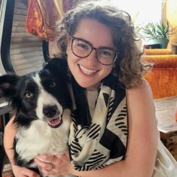Person with curly hair holding a black and white dog indoors near a couch, chair, and plants.