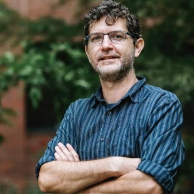 Person with curly hair in a blue and black striped shirt standing outdoors near greenery and a brick wall.