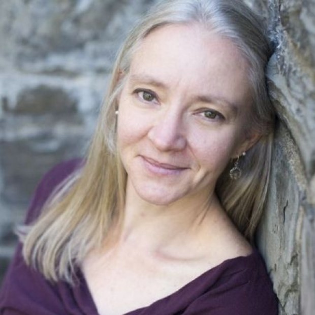 Person with long blonde hair in a dark purple top leaning against a stone wall.
