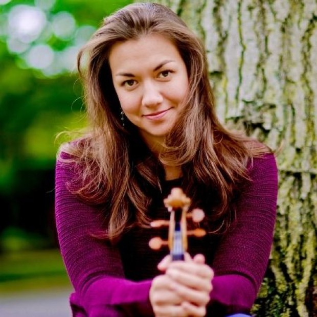 Person with long hair in a purple sweater holding a violin while sitting against a tree, with greenery in the background.