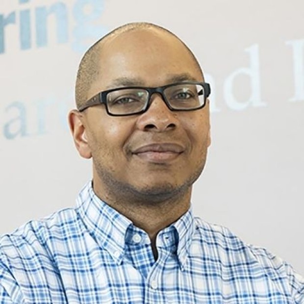 Person in glasses and a blue checkered shirt standing in front of a background with partial text.