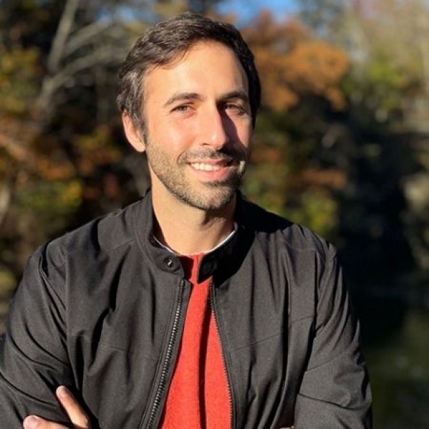Person in a black jacket and red shirt standing outdoors near autumn trees and water.