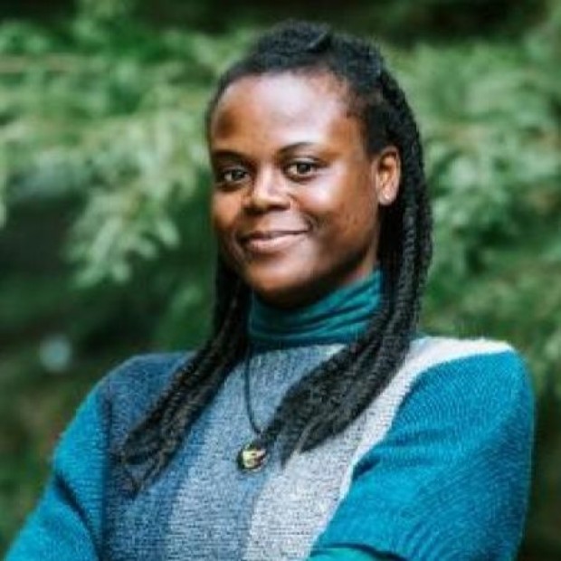 Black woman with braided hair in a blue and gray sweater, wearing a round pendant necklace, standing in front of green foliage.