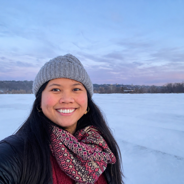 A selfie of a smiling woman wearing a knitted cap and scarf standing in an expansive winter landscape.