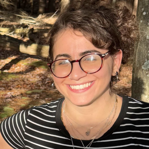 A casual photograph of a smiling woman with medium-light skin and glasses. She standing outside in sunlight, wearing a striped top and multiple necklaces.