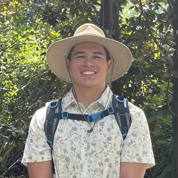 Casual outdoor photo of a smiling young man wearing a short sleeved shirt and wide brimmed hat.