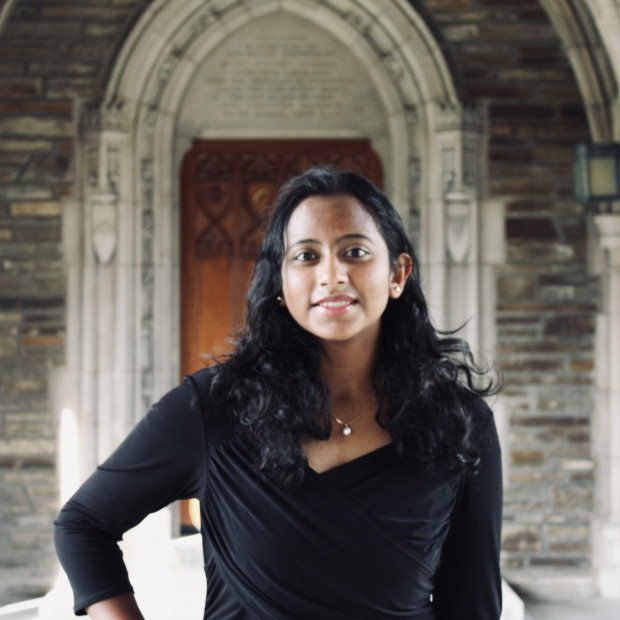 A photograph of a woman with medium-brown skin and past shoulder length wavy black hair, smiling. She is wearing a necklace, and a black dress and jacket.. 