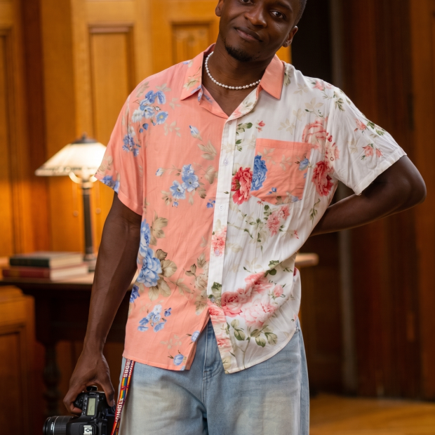 wearing pink and white flower patterned shirt in a wood paneled room, holding a camera 