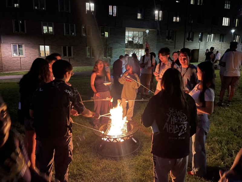 A nighttime outdoor gathering with a group of people standing in a circle around a fire pit. The fire casts a warm glow on their faces and bodies. Some individuals are holding sticks with marshmallows or other items, roasting them over the flames. The setting appears to be a grassy lawn with a building in the background, its windows lit from within. Additional groups of people are visible further back, indicating a larger social event or community gathering.
