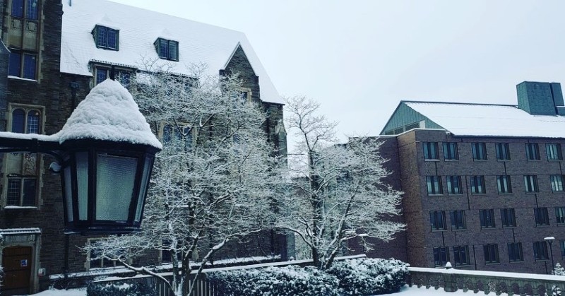 A serene winter scene featuring two buildings surrounded by snow-covered trees and bushes. On the left, there is a traditional stone building with a steeply pitched roof, dormer windows, and a large lantern hanging from its side. On the right, a more modern building stands with a flat roof and rectangular windows. Snow blankets the rooftops, trees, and all visible surfaces, creating a peaceful, wintry atmosphere.